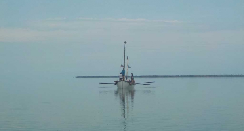 A sailboat floats in calm water. Fog appears to be rolling in or out. 